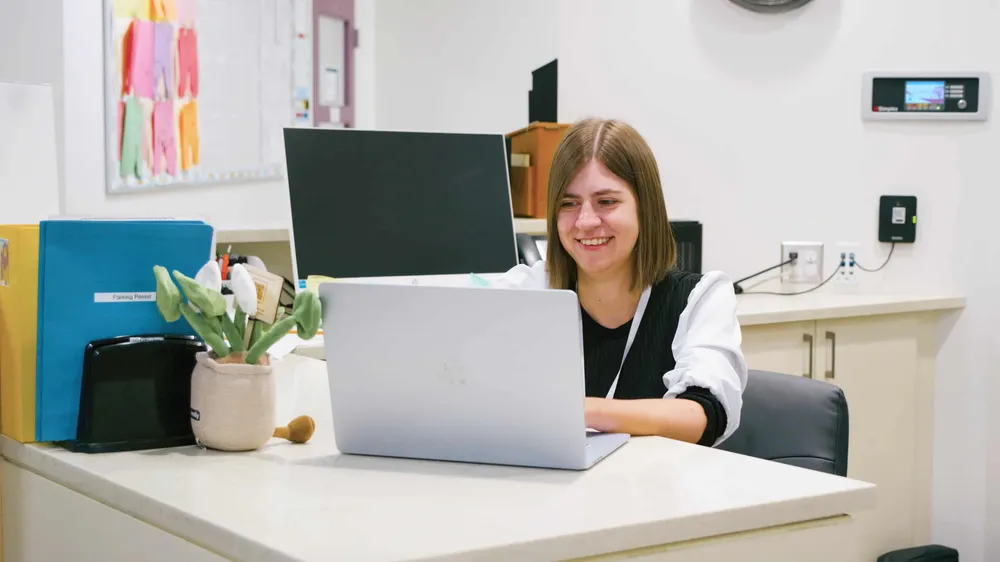 A smiling administrator working on a laptop at the front office desk
