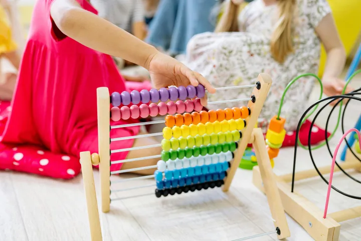 A child counting beads on a rainbow abacus while classmates play nearby