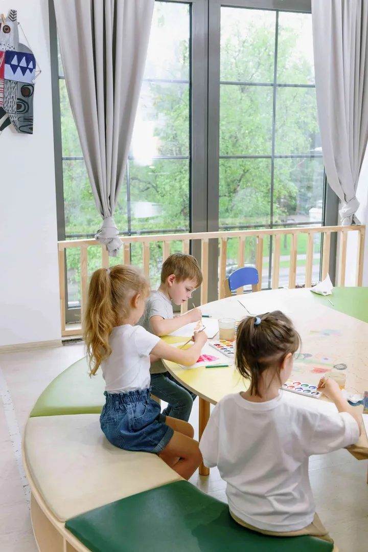 Children painting at a round table beside a sunny window