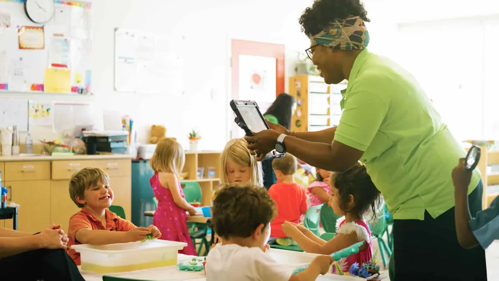 An educator holding a tablet beside children playing at a sensory table