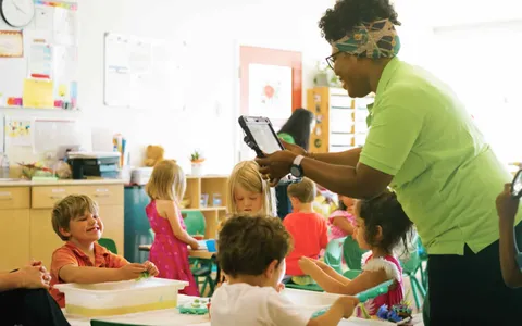 An educator holding a tablet beside children playing at a sensory table