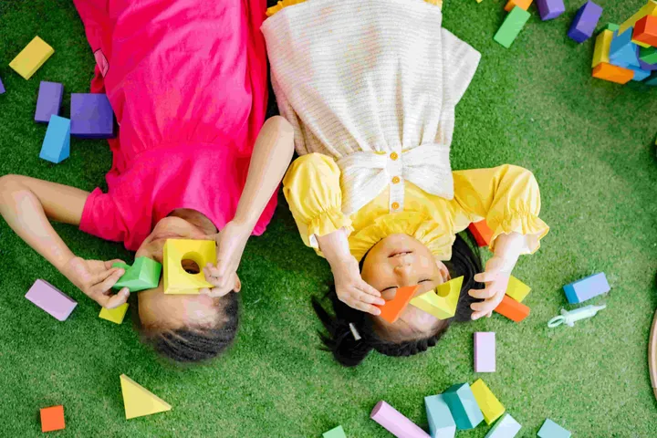 Two girls lying on turf holding colourful foam blocks up to their eyes
